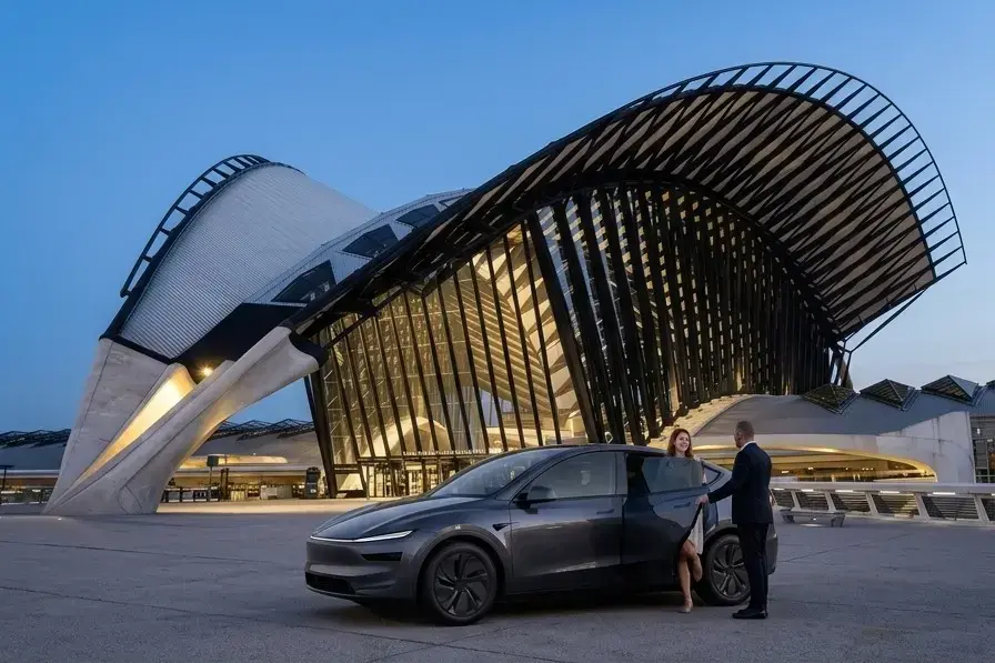 Tesla Model Y gris foncé devant la gare de Lyon Saint-Exupéry au crépuscule : un chauffeur en costume ouvre la portière à une passagère